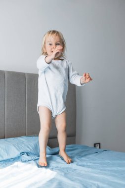 Sarışın bir çocuk heyecanla yatağına atlıyor, evde eğleniyor. Yumuşak sabah ışığı, onun keyifli ifadesini ve dalgalı saçlarını vurgular, sıcak ve neşeli bir atmosfer yaratır..