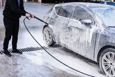 Bir adam yıkama istasyonundaki bir arabaya köpük sürmek için yüksek basınçlı çamaşır makinesi kullanıyor. Ayar parlak ve güneşli, temizlik sürecini vurguluyor. Otomobil yıkama işlemi