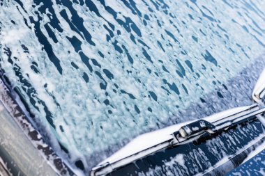 Köpük ve köpük birikerken, parlak güneş ışığı sabunlu bir arabanın ön camından yansıyor. Garaj yolu huzurlu bir atmosferle çevrilidir. Bu da öğleden sonra yıkanma etkinliğinin göstergesidir..