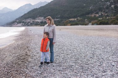 Bir anne ve küçük oğlu kışın bir plajı keşfederler. Çocuk neşeli bir şekilde ayakta dururken anne arka plandaki su ve dağların huzurlu manzarasına gülümsüyor..