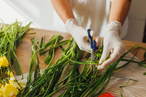Eldivenli bir insan ahşap bir yüzeyde yeşil otları makasla kesiyor. Renkli çiçekler yakında, parlak çiçekçi dükkanının neşeli atmosferini güçlendiriyor..