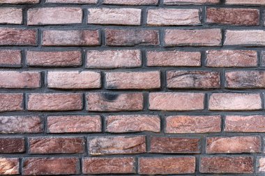 Türlü kırmızı ve kahverengi tuğlalar sergilenen desenli tuğla duvarın yakın plan görüntüsü. Düzenleme, şehir ortamında bulunan pürüzlü yüzeyleri ve farklı renkleri vurgular.