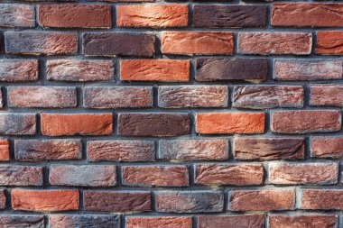Türlü kırmızı ve kahverengi tuğlalar sergilenen desenli tuğla duvarın yakın plan görüntüsü. Düzenleme, şehir ortamında bulunan pürüzlü yüzeyleri ve farklı renkleri vurgular.