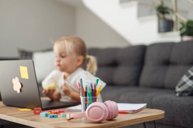 Genç sarışın Avrupalı kız masada dizüstü bilgisayar kullanıyor renkli kalemler ve kulaklıklarla modern oturma odasında gri kanepeli ve merdivenli..