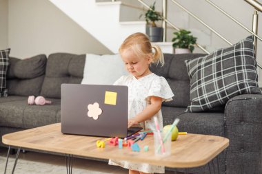 Genç sarışın Avrupalı kız masada dizüstü bilgisayar kullanıyor modern oturma odasında gri kanepeli ve merdivenli renkli kalemlerle..