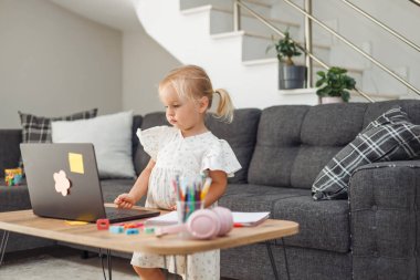 Genç sarışın Avrupalı kız masada dizüstü bilgisayar kullanıyor renkli kalemler ve kulaklıklarla modern oturma odasında gri kanepeli ve merdivenli..