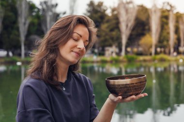 Uzun kahverengi saçlı beyaz bir kadın dingin bir gölün yanında ahşap bir kase tutuyor. Arka planda ağaçlar ve yeşillikler görülebilir..