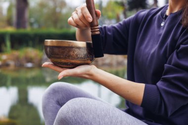 Kahverengi saçlı genç bir kadın, sakin bir su kütlesinin yanında açık havada şarkı söylüyor. Koyu renk bir kazak ve gri tayt giyer. Rahatlamayı ve farkındalığı teşvik eder..