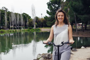 Uzun kahverengi saçlı beyaz bir kadın ağaçlarla çevrili sakin bir göl kenarında elektrikli scooter kullanırken gülümsüyor..