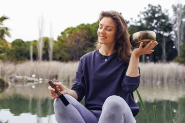 Kahverengi saçlı beyaz bir kadın dingin bir gölün kenarında elinde renkli malzemelerle dolu ahşap bir kâse tutuyor. Suyu çevreleyen yeşil sazlıklar.