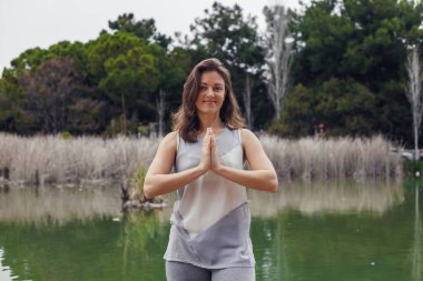Kahverengi saçlı, genç beyaz bir kadın dingin bir gölün kenarında duruyor ve elleri dua pozisyonunda yoga yapıyor. Yeşillik her yeri sarmış..