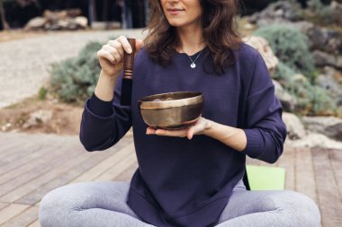 Kahverengi saçlı beyaz bir kadın açık havada yoga minderinde bacak bacak üstüne atmış oturuyor. Yeşillik ve ahşap süslemelerle çevrili bir şarkı kasesi kullanıyor..