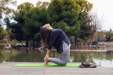 Kahverengi saçlı beyaz bir kadın ağaçlarla çevrili sakin bir gölün kenarındaki yeşil bir minderde yoga yapıyor. Derin bir poz veriyor, nefesine odaklanıyor..