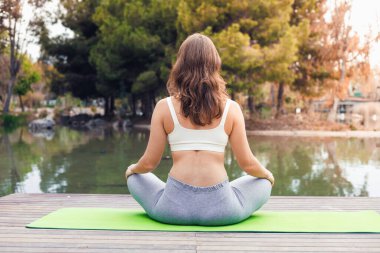 Uzun kahverengi saçlı beyaz bir kadın dingin bir gölün kenarındaki yeşil yoga minderinin üzerinde oturur ve doğal ortamında meditasyon yapar..