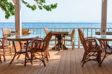 Aydınlık ve güneşli bir günde berrak mavi suya bakan deniz kenarındaki terasa Rattan sandalyeler ve masalar.