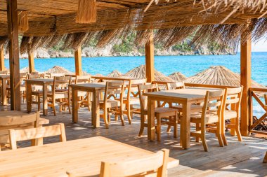 Büyüleyici bir sahil yemek alanı, açık mavi deniz manzarası sağlayan ahşap masalar ve güneş şemsiyeleri içerir.