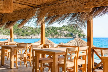 Büyüleyici bir sahil yemek alanı, açık mavi deniz manzarası sağlayan ahşap masalar ve güneş şemsiyeleri içerir..