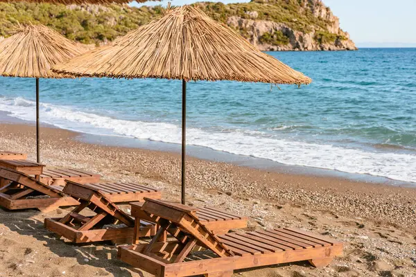 Tahta şezlonglar, kumsaldaki saman şemsiyelerin altında sıralanmış, kıyıdaki rahatlamaya davetiye çıkarıyorlar..