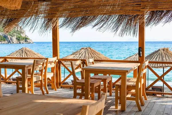 Büyüleyici bir sahil yemek alanı, açık mavi deniz manzarası sağlayan ahşap masalar ve güneş şemsiyeleri içerir.