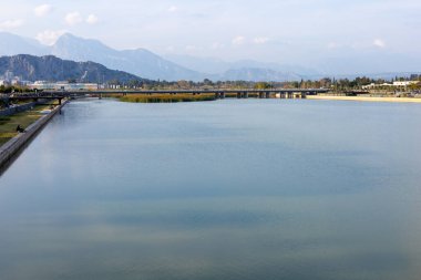Sakin bir nehir mavi gökyüzünü ve yakındaki dağları yansıtır. Bereketli yeşillik kıyıları çevreleyen, suları aşan bir köprüyle, Antalya şehrinde huzurlu bir manzara yaratıyor.