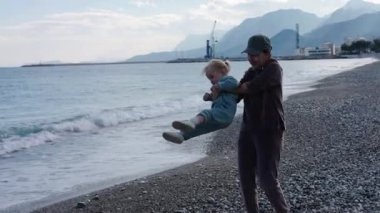 Bir çocuk, açık bir gökyüzünün altındaki kayalık bir plajda bir ebeveyn tarafından neşeyle kaldırılırken neşe yaşar..