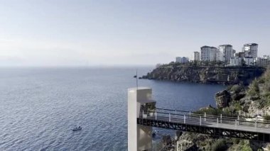 Arka planda modern binalar olan kayalık bir kıyı şeridi manzarası. Açık mavi gökyüzü ve sakin deniz sakin bir atmosfer yaratır..