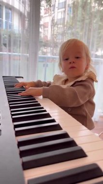 Küçük bir kız klavye çalıyor, parmakları tuşların üzerinde dans ediyor. Mekan doğal ışıkla dolu, çocukların konsantrasyonunu ve rahat müziğin tadını vurguluyor.