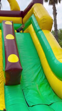 Çocuk parkında renkli bir şişirilebilir kaydırak. Slaytta palmiye ağaçlarıyla çevrili parlak sarı, yeşil ve kahverengi renkler yer alıyor. Yüksek kalite 4k görüntü