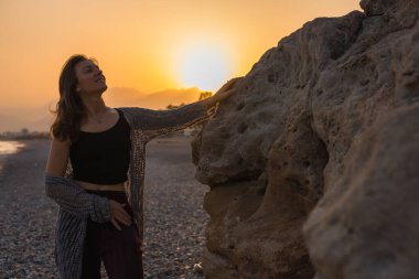 Uzun kahverengi saçlı beyaz bir kadın gün batımında kayalık bir sahilde duruyor. Siyah bir bluz ve kestane rengi pantolon giyiyor, sakin atmosferin tadını çıkarıyor..