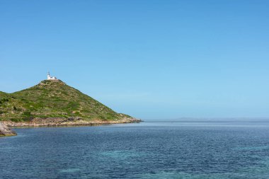 Türkiye 'de yeşil bir tepenin sakin kıyı manzarası, tepesinde bir deniz feneri, berrak mavi bir gökyüzü ve sakin suların altında.
