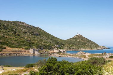 Türkiye 'de manzaralı bir kıyı manzarası. Sakin mavi bir denizin üzerinde yeşil tepeler yükseliyor, küçük bir liman ve berrak bir gökyüzünün altında görünen uzak deniz feneriyle.