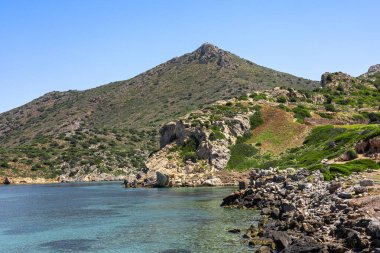 Türkiye 'de turkuaz suyu ve yeşil yamaçlı kayalık bir sahil şeridi. Açık mavi gökyüzünün altında bir dağ yükseliyor..
