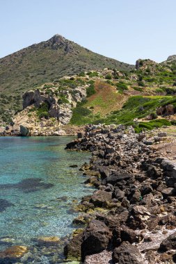 Türkiye 'de turkuaz suyu ve yeşil yamaçlı kayalık bir sahil şeridi. Açık mavi gökyüzünün altında bir dağ yükseliyor..