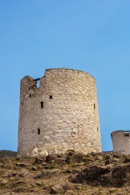 Açık mavi gökyüzünün altındaki bir yamaçta tarihi bir taş kule duruyor. Kaya arazisi ve uzak dağlarla çevrili, pürüzlü bir dokusu olan silindirik bir yapı..