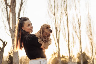 Uzun kahverengi saçlı genç bir Asyalı kadın kucağında küçük altın bir cocker spaniel tutuyor. Arka planda uzun ağaçlar ve sıcak bir günbatımı parıltısı var..