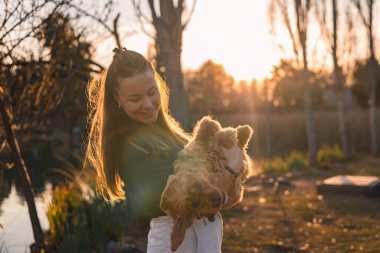 Uzun kahverengi saçlı beyaz bir kadın, açık havada güneşli bir ortamda altın renkli bir spanyel tutuyor. Arka planda ağaçlar ve bir gölet görülebilir..