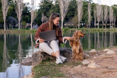 Uzun kahverengi saçlı beyaz bir kadın göl kenarında dizüstü bilgisayar kullanarak oturuyor. Altından bir cocker spaniel onun yanında duruyor, neşeyle etkileşime geçiyor..