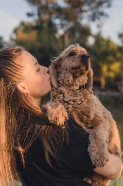 Uzun kahverengi saçlı beyaz bir kadın, açık kahverengi bir cocker spanyel köpeği tutuyor, açık havada sevgi dolu bir anı paylaşıyor..