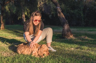 Uzun kahverengi saçlı ve güneş gözlüklü beyaz bir kadın gün batımında çimenli bir parkta küçük, altın renkli bir köpekle etkileşime geçiyor..