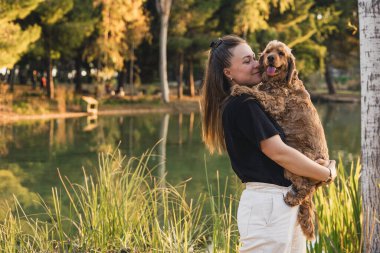 Uzun kahverengi saçlı beyaz bir kadın ağaçlarla çevrili bir gölün kenarında küçük kahverengi bir cocker spaniel tutuyor. Sahne huzurlu ve güneşli..