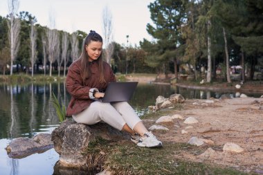 Uzun siyah saçlı genç bir İspanyol kadın göl kenarındaki bir kayanın üzerinde oturuyor ve dizüstü bilgisayarla çalışıyor. Arka planda ağaçlar ve su görülebilir..