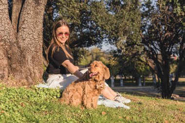 Uzun kahverengi saçlı, beyaz bir kadın parktaki bir ağacın altında altın renkli bir köpeğin yanında çimlerin üzerinde oturuyor..