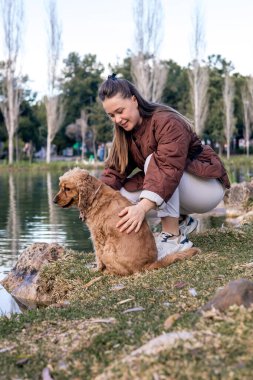 Uzun kahverengi saçlı, beyaz bir kadın gölün kenarında oturuyor ve altın renkli bir cocker köpeğini okşuyor. Köpeğin dalgalı bir paltosu var ve çimlerin üzerinde oturuyor..