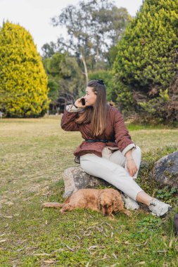 Uzun kahverengi saçlı, genç bir İspanyol kadın parkta bir kayanın üzerinde oturmuş, telefonuyla konuşuyor. Küçük altın bir cocker spaniel çimlerin üzerinde onun yanında yatıyor..