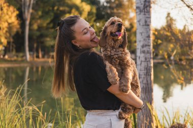 Uzun kahverengi saçlı beyaz bir kadın ağaçlarla çevrili bir gölün kenarında kahverengi bir köpek tutuyor. Kadın gülüyor ve şakacı bir şekilde dilini çıkarıyor..
