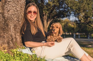 Kafkasyalı, uzun kahverengi saçlı genç bir kadın, elinde küçük altın bir cocker spaniel ile bir ağacın altında oturuyor. Sahne güneşli ve rahat, aralarındaki bağı gösteriyor..