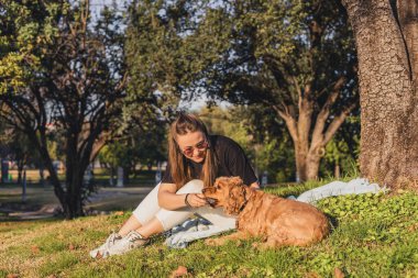 Uzun kahverengi saçlı beyaz bir kadın çimenlerde oturur, ağaçlarla çevrili güneşli bir parkta altın renkli bir köpekle oynar..