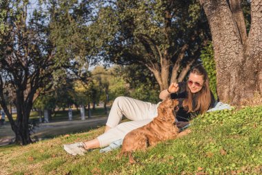 Uzun kahverengi saçlı beyaz bir kadın çimlerin üzerinde oturur, parkta küçük altın renkli bir köpek yavrusuyla oynar..