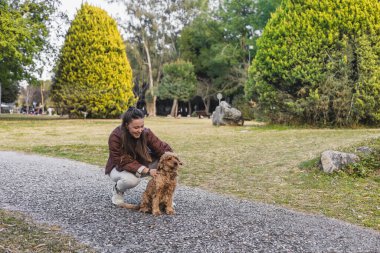 Uzun kahverengi saçlı genç bir kadın parkta küçük, altın renkli bir köpeğin yanında diz çöker. Çevrelerinde yemyeşil ağaçlar var, sakin bir açık hava ortamı yaratıyorlar..