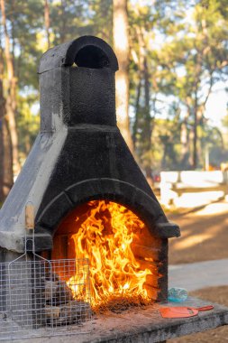 Bacadan yükselen alevlerle dolu taştan bir şömine. Ortam ağaçlarla çevrili. Toplanma için doğal bir atmosfer yaratıyor..